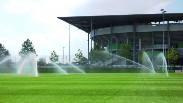 Automatisierte Sportplatzbewässerung vor einem modernen Stadion mit gleichmäßigen Wasserstrahlen auf einem gepflegten Rasen